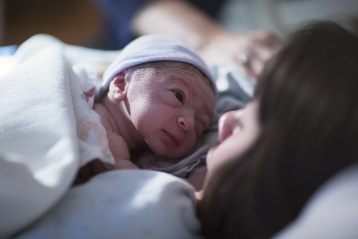 A newborn mixed race asian caucasian in a blue cap baby rest on his brunette asian mothers chest and stares into her eyes for the first time.