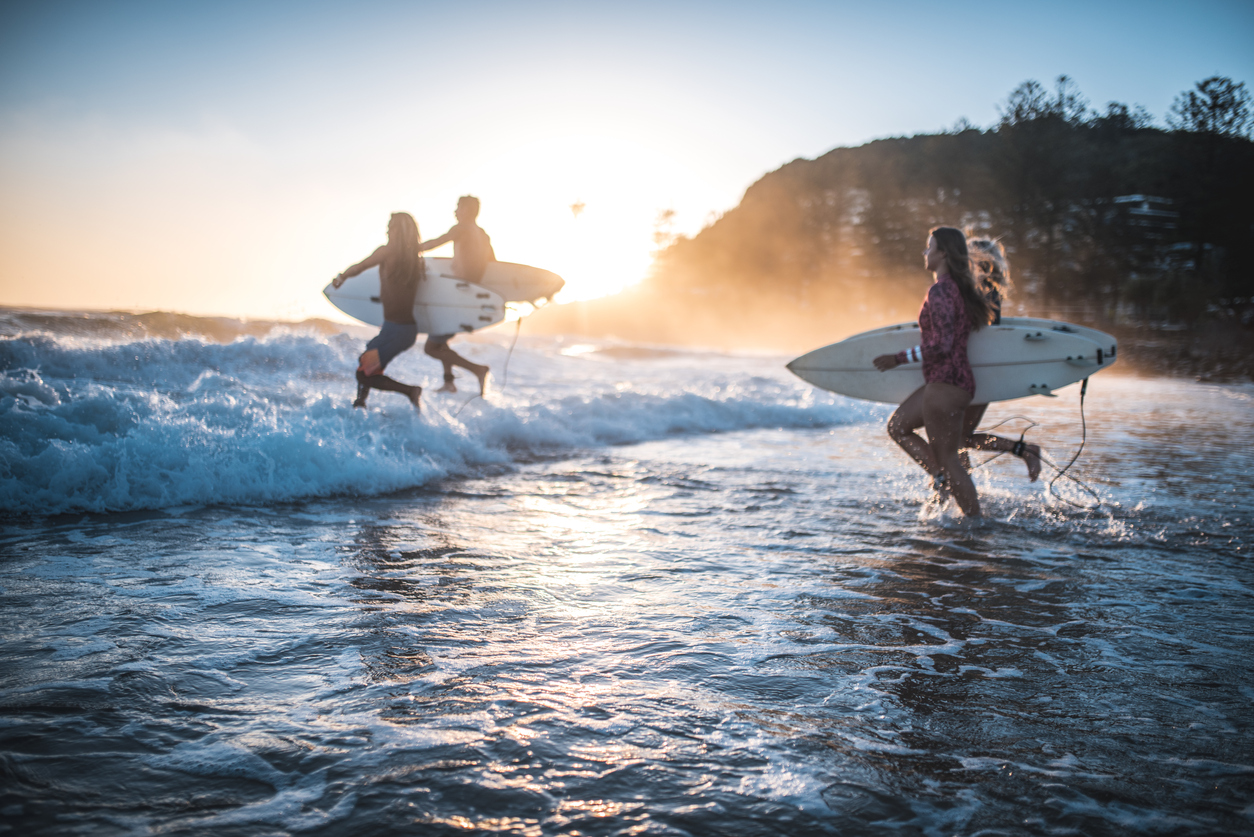 Four friends, surfers, running into the water early in the morning with surfboards in their hands. Sun is rising in back.