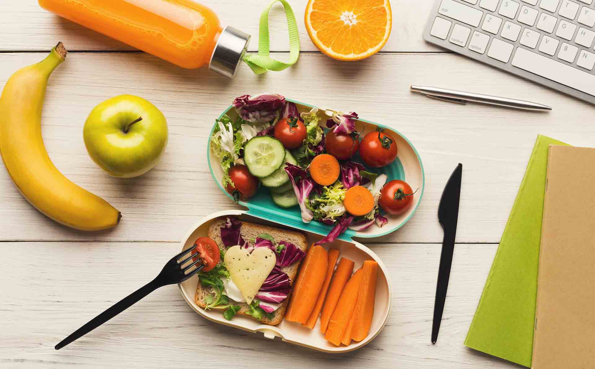 Healthy snack at office workplace. Eating organic vegan meals from take away lunch box at wooden working table with computer keyboard, top view