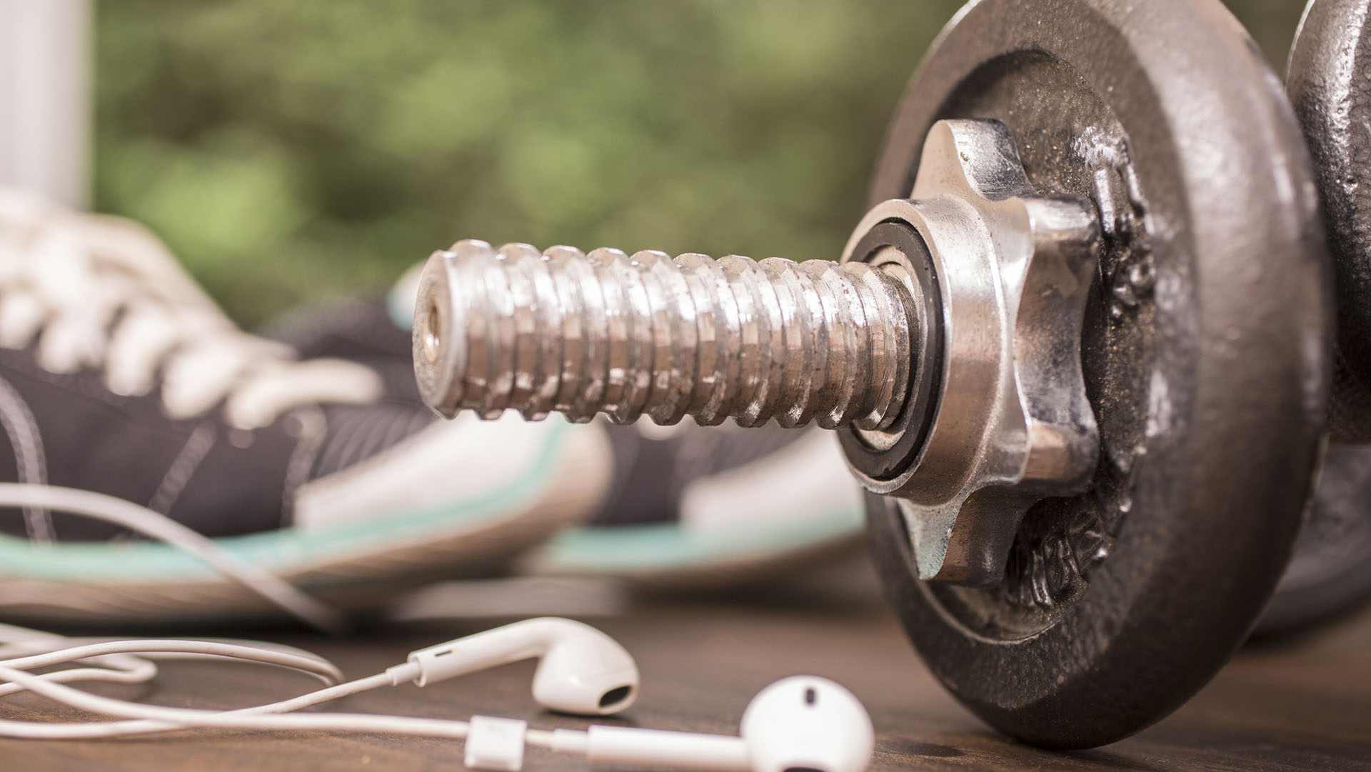 Fitness, exercise themed scene with barbells, sneakers, earbuds.  Group of objects lie on wooden surface in gym or health club setting with outdoors view in backgorund. Listening to music while working out.
