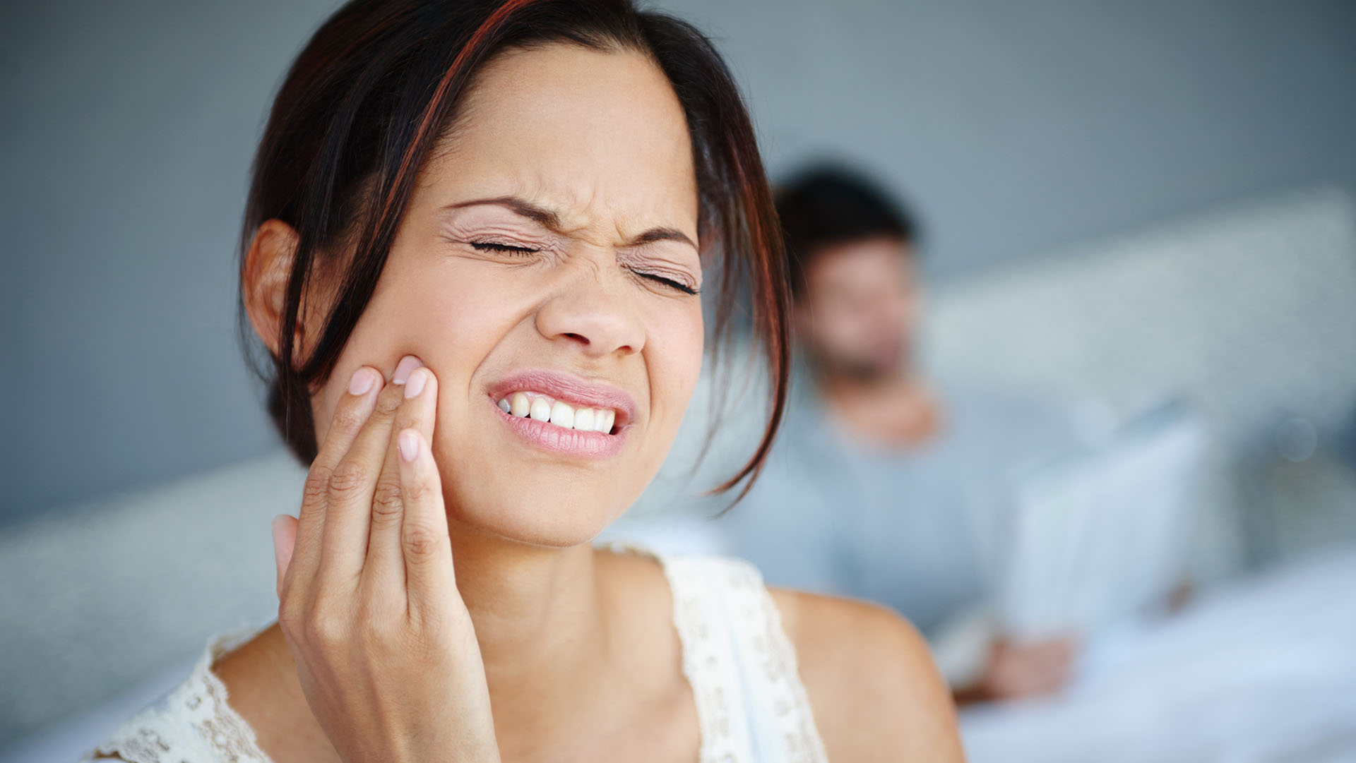 Shot of a woman sitting on the side of her bed with bad toothache with her boyfriend in the background