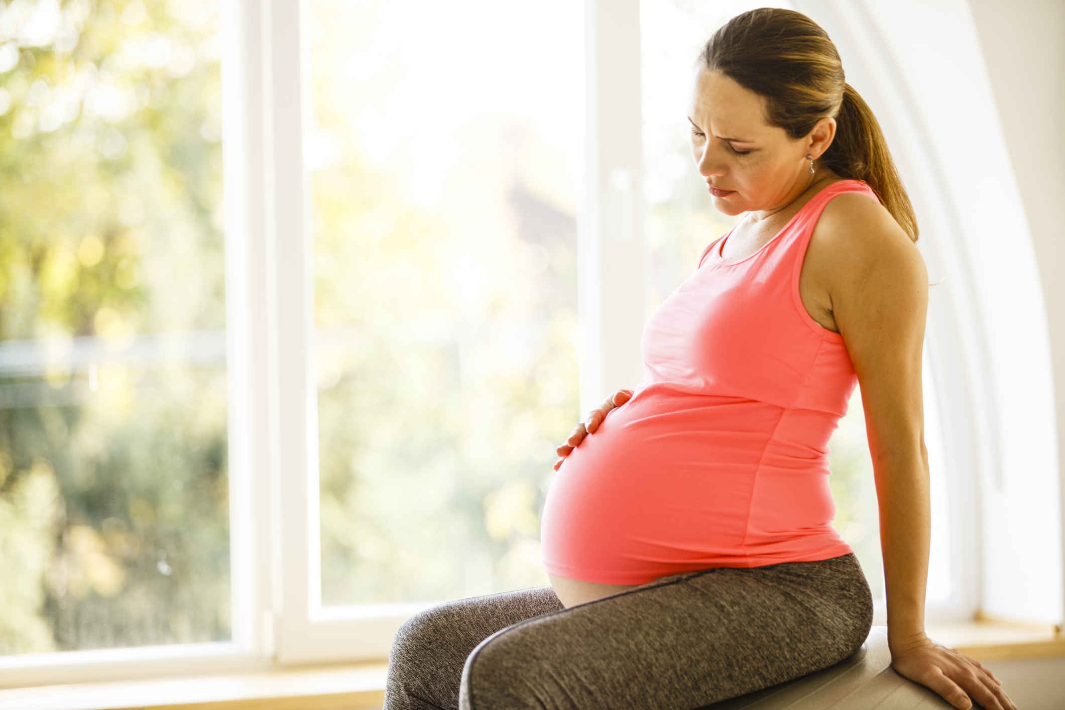 Mid adult pregnant woman is touching her stomach, holding her arm on a fitness ball and feeling a bit pain and sitting on a fitness ball near the window at home