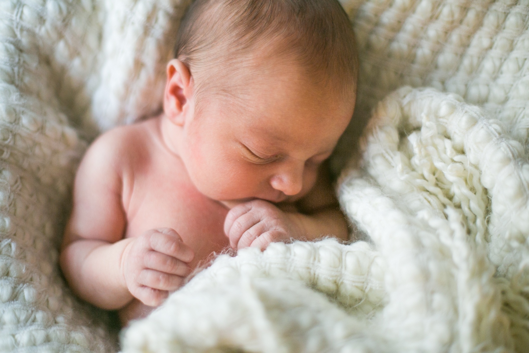 A brand new one week old baby boy sleeps wrapped in a white fuzzy blanket. He is peaceful and his hand rests near his mouth.