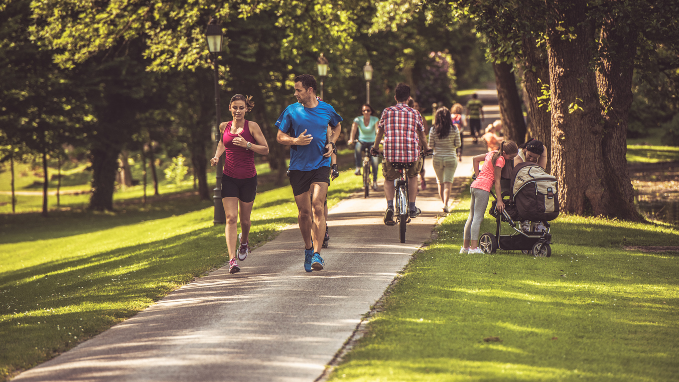 A lot of people in the park, some of them are jogging, some walking, cycling or standing.