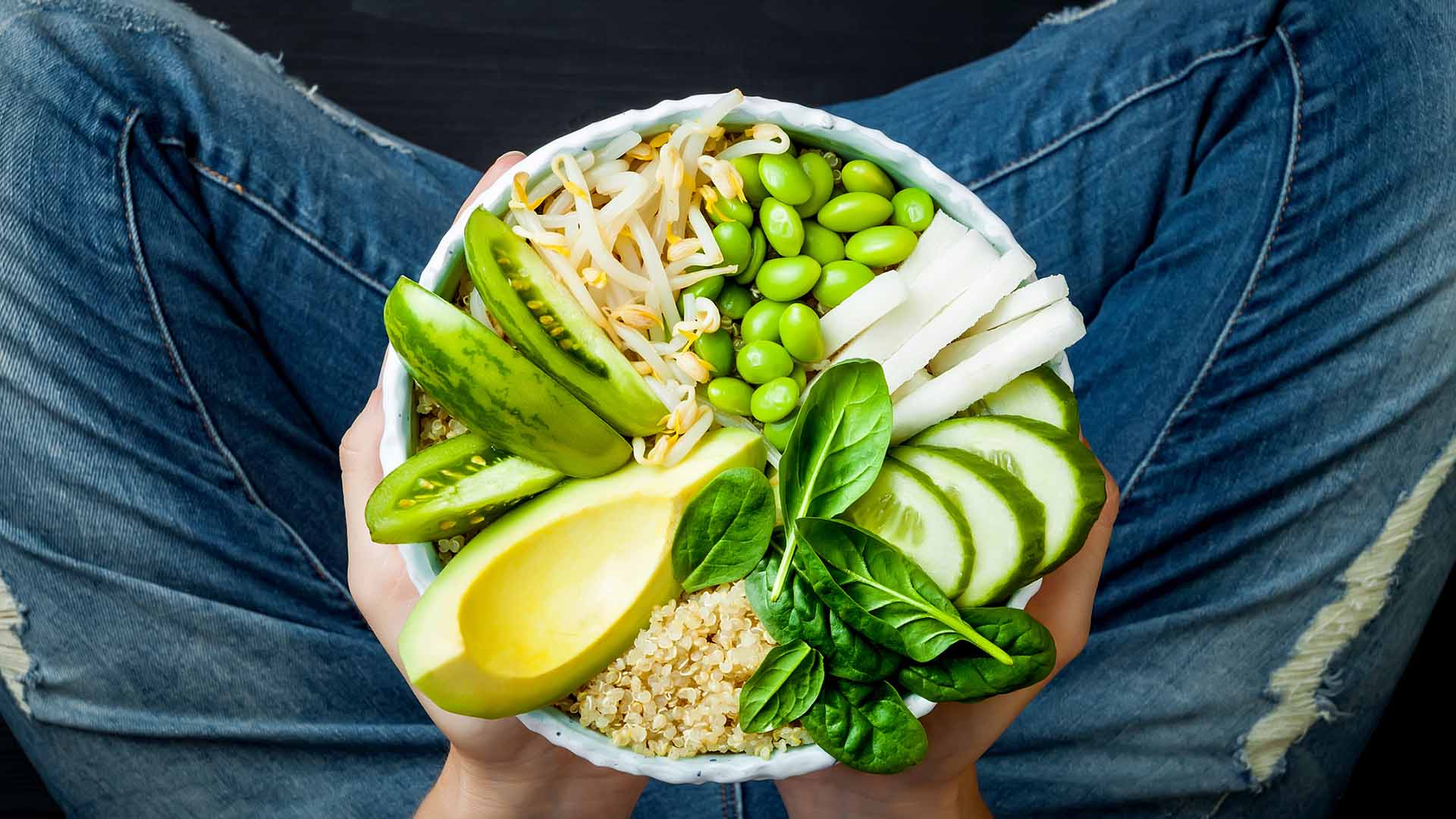 Girl in jeans holding vegan, detox green Buddha bowl with quinoa, avocado, cucumber, spinach, tomatoes, mung bean sprouts, edamame beans, daikon radish. Top view, overhead