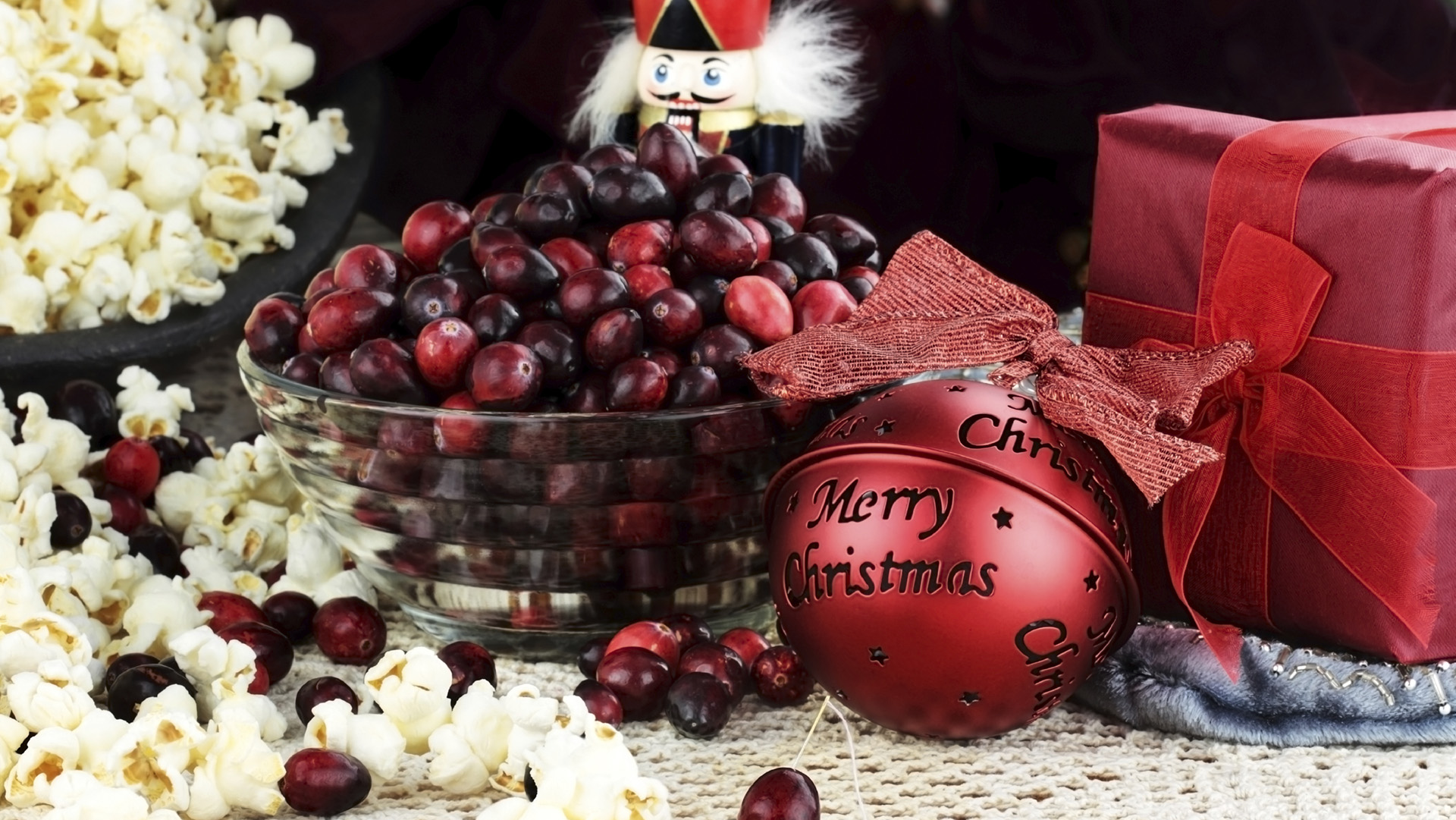 String of popcorn and cranberries with bowl of cranberries, popcorn, gift and ornaments in background. Shallow depth of field.