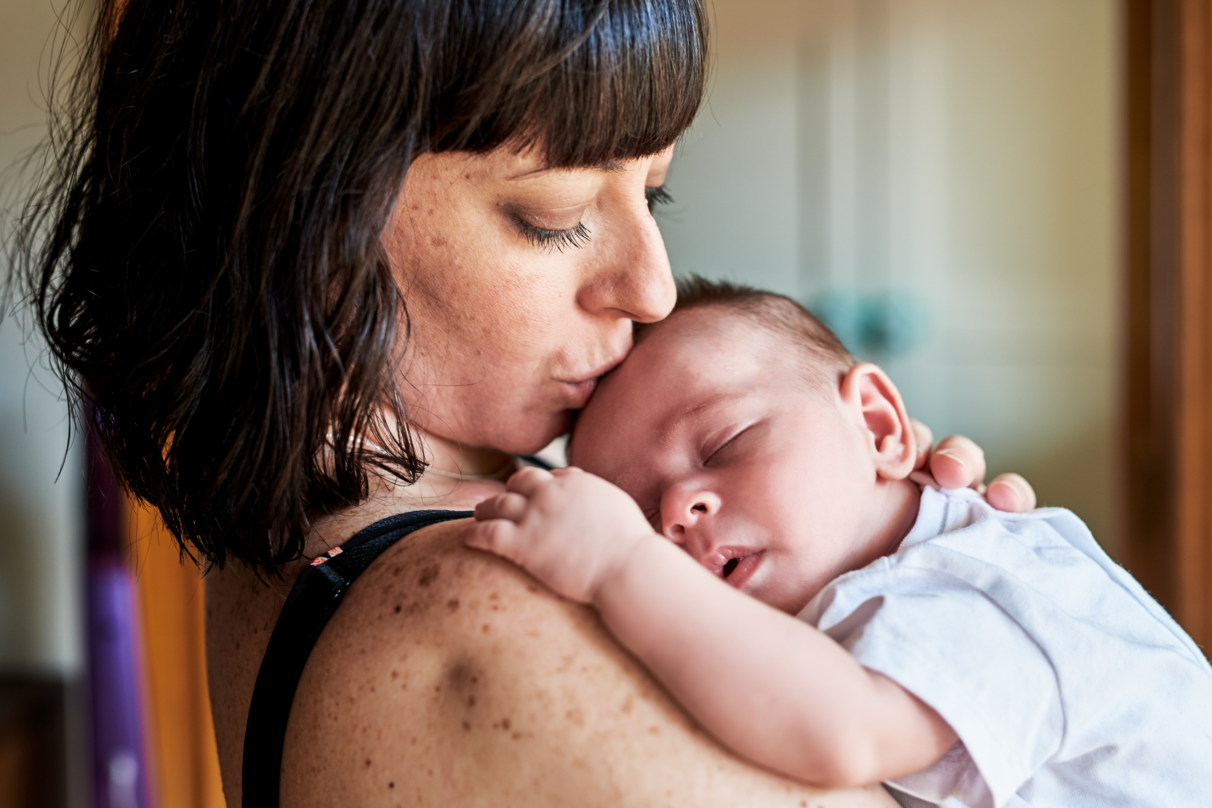 Side view of brunette mum kissing her adorable sleeping newborn in forehead