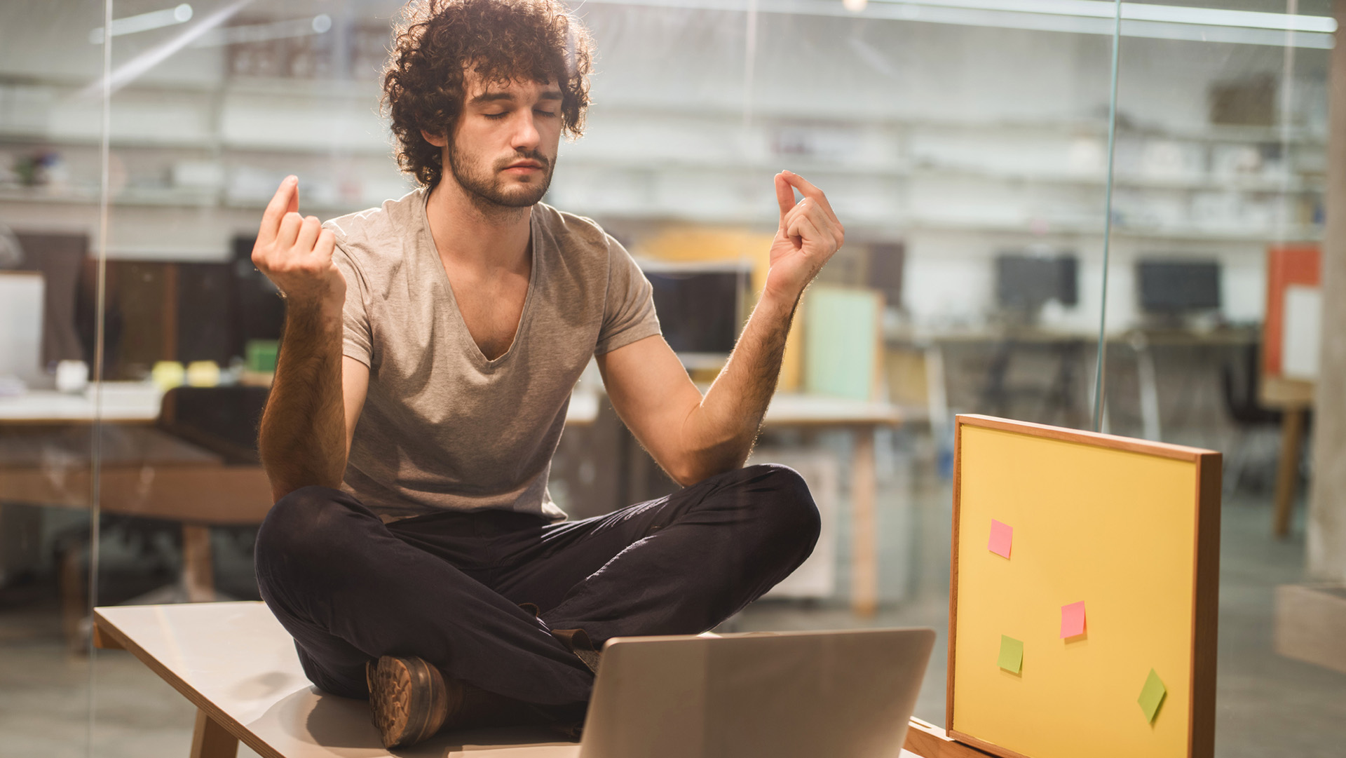 Young man sitting in the lotus position on table in the office. He is relaxing from work and meditating with his eyes closed. The view is through the glass.