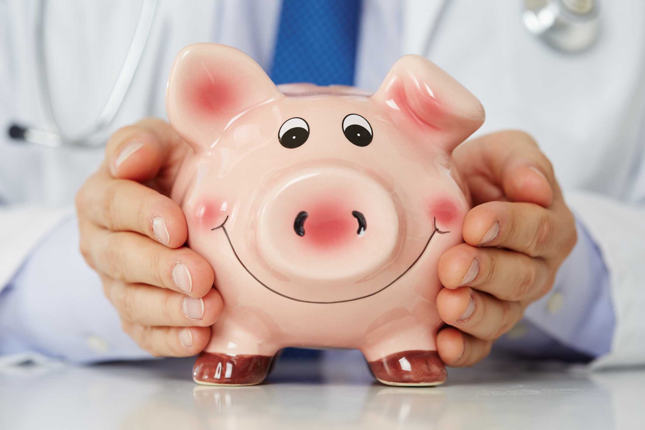 Male medicine doctor wearing blue tie holding and covering happy funny smiling piggybank in hands closeup. Medical service economy, health care savings and insurance concept. Focus on piggy bank