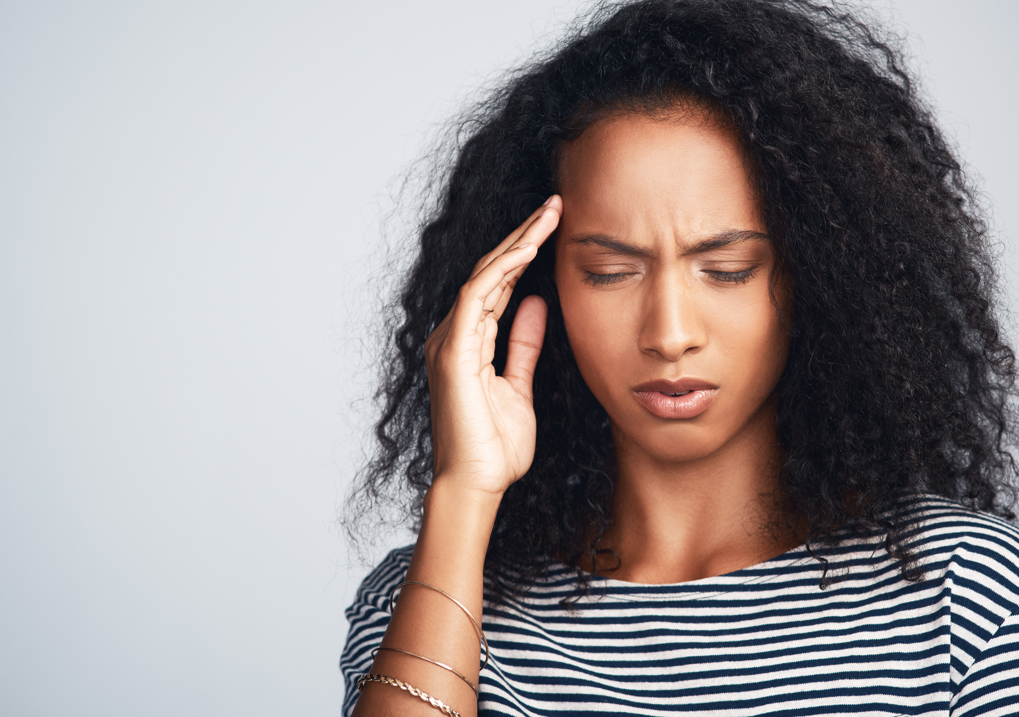 Studio shot of a uncomfortable looking young woman holding her head due to a migraine  while standing against a grey background