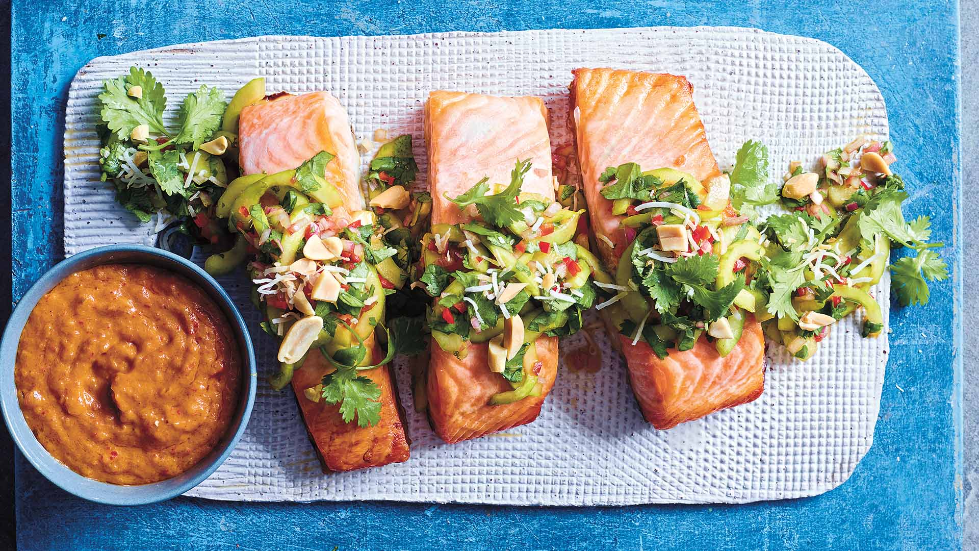 Grilled satay salmon fillets topped with a crunchy cucumber and herb salad, presented on a textured blue mat, accompanied by a bowl of rich, spicy satay sauce on the side, ready to be drizzled over the fish.