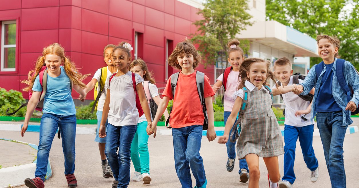 Happy kids with rucksacks walking holding hands near school building during summer day time