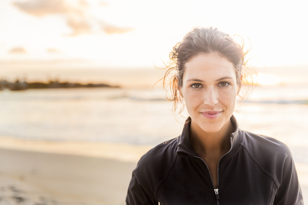 A photo of fit young woman with confident look on her face. Portrait of happy female athlete is at beach. She is wearing sportswear during sunset.