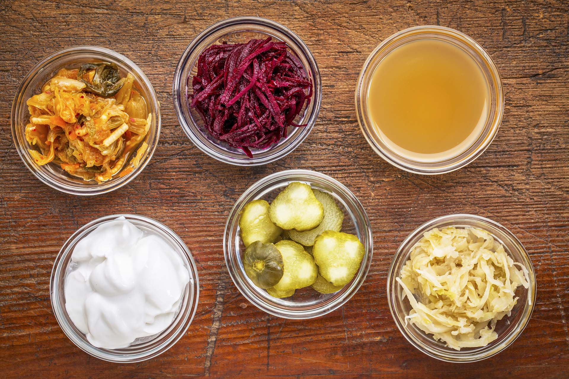 a set of fermented food great for gut health - top view of glass bowls against wood:  kimchi, red beets, apple cider vinegar, coconut milk yogurt, cucumber pickles, sauerkraut