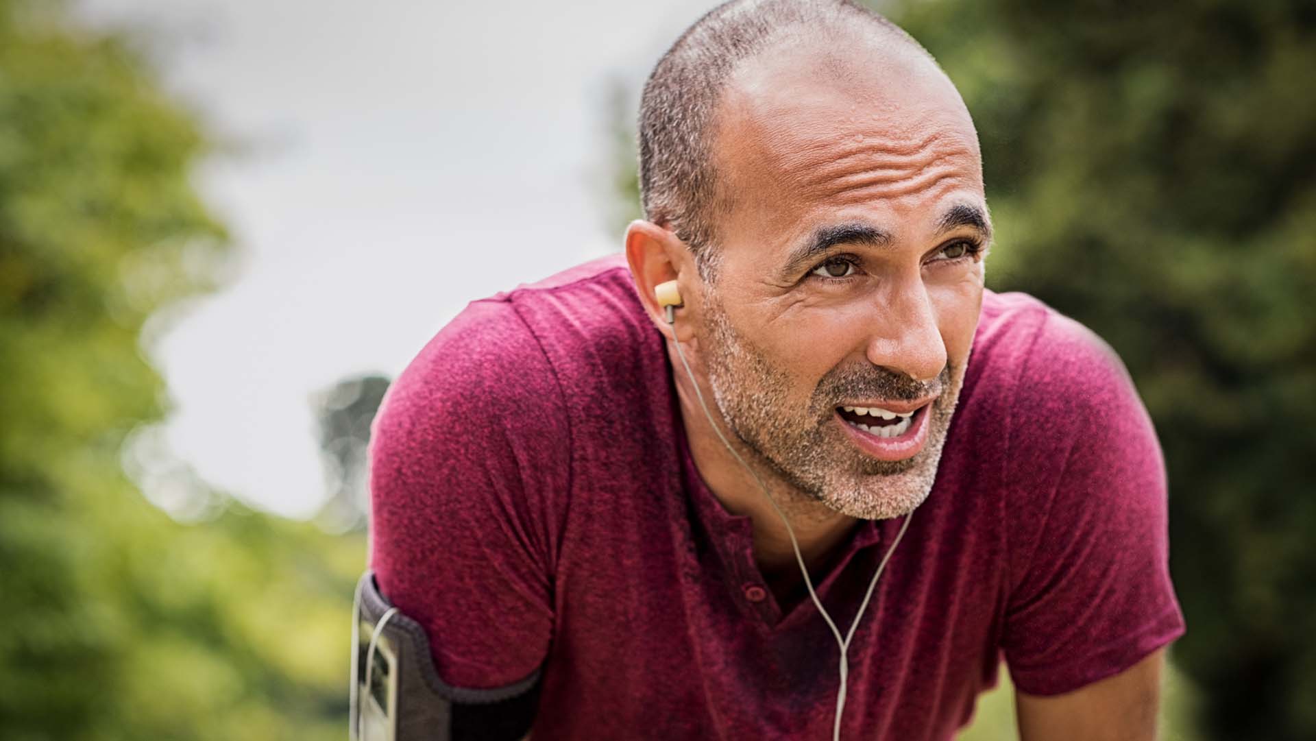 Portrait of athletic mature man after run. Handsome senior man resting after jog at the park on a sunny day. Sweaty multiethnic man listening to music while jogging.