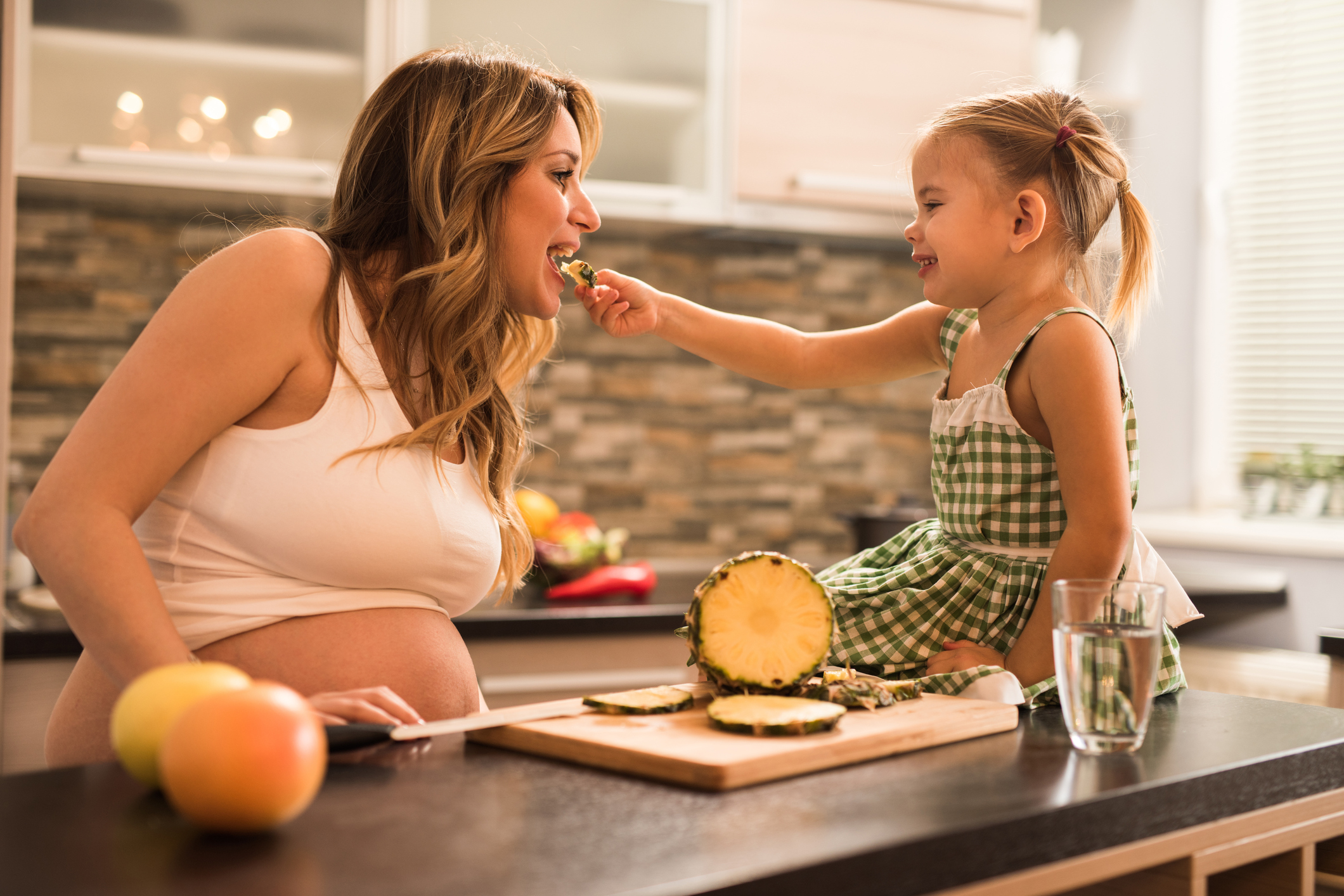 Cute little girl feeding her expecting mother with fresh pineapple in the kitchen.