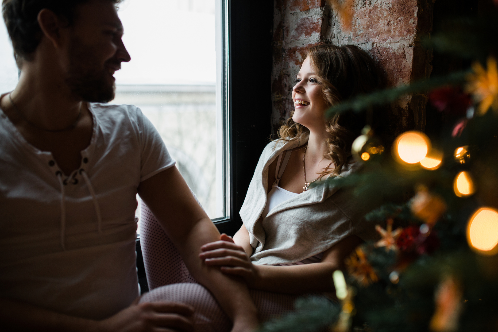 Happy couple of lovers in pajamas sit on the windowsill. Christmas atmosphere and new year gift at home. Young family together. Garland lights in the foreground bokeh.