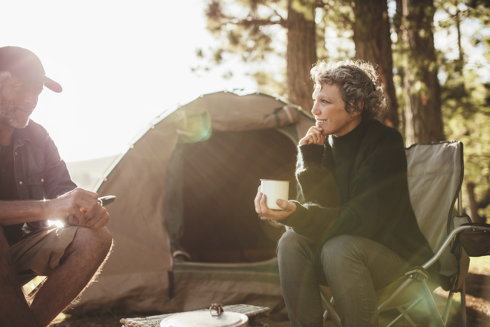 Portrait of relaxed mid adult couple sitting outside tent while camping nearby lakeside on a sunny day. Woman holding coffee talking with man.