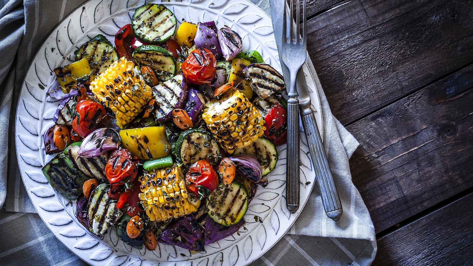 Close-up view of a grilled vegetables plate shot from above on rustic wooden kitchen table. The plate is on a gray textile napkin at the left of an horizontal frame and a fork and knife are at the right of the plate. DSRL studio photo taken with Canon EOS 5D Mk II and Canon EF 100mm f/2.8L Macro IS USM