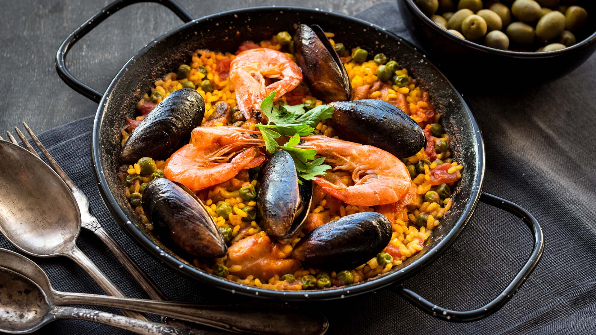 Paella in black pan with rice, shrimps, mussels, squid and meat, bowl with olives and vintage cutlery. Seafood paella, traditional spanish dish. Paella on rustic black wooden table. Selective focus