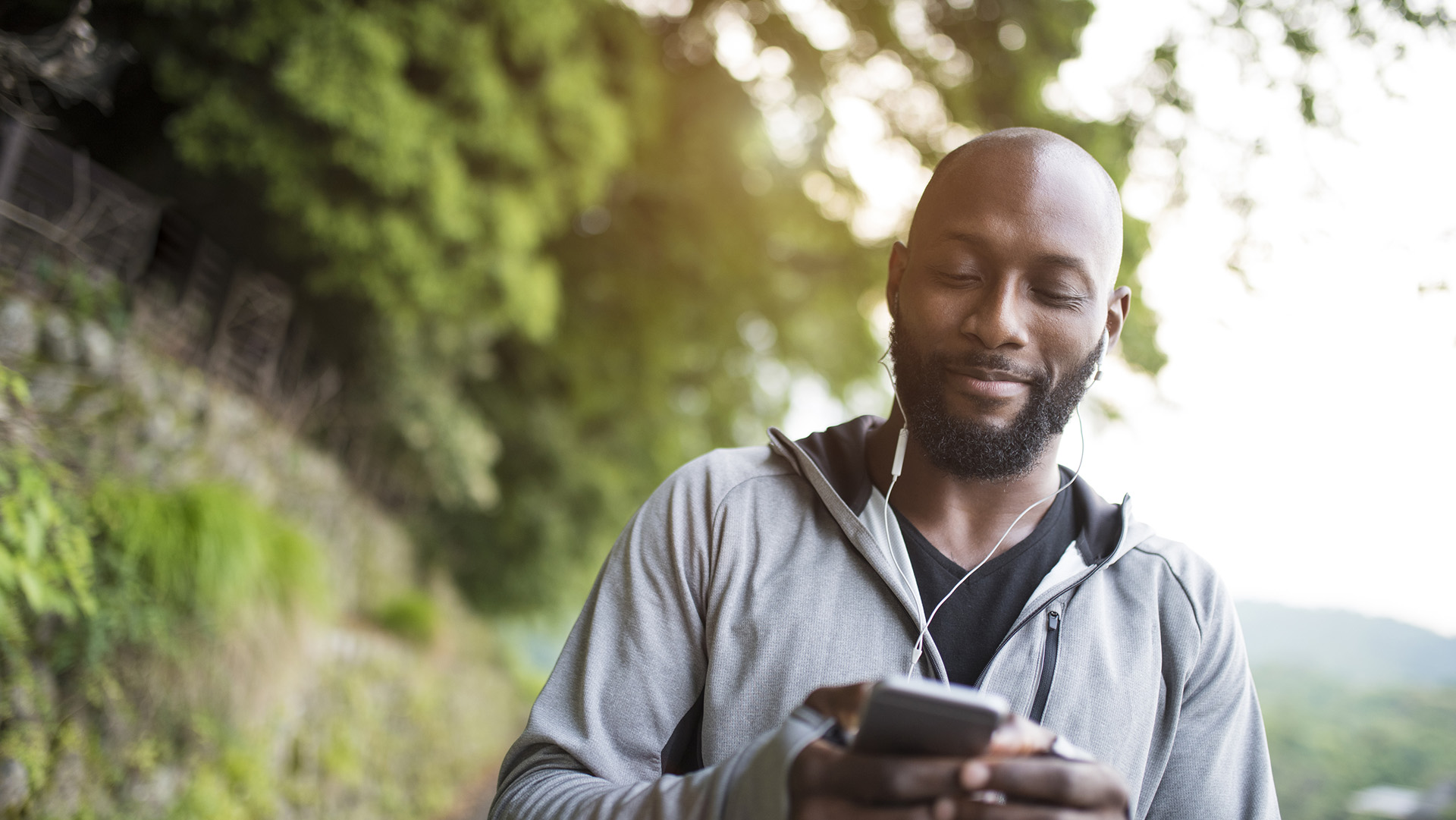 Portrait of young black man in Kyoto, Japan. He is holding mobile phone and has earphones. He is looking on his mobile phone. Green trees in the back.