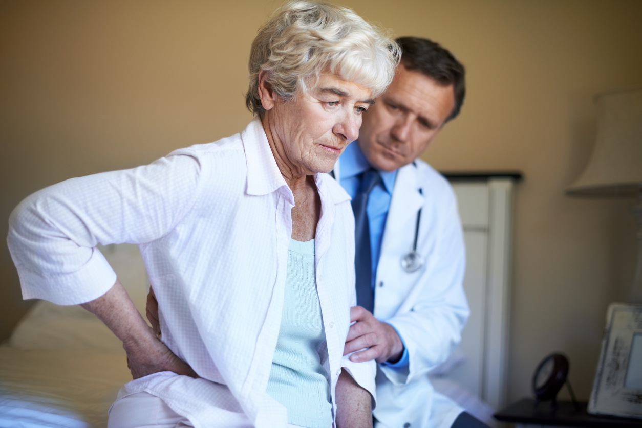 "Profile of a senior woman, sitting on her bed and holding her back in pain while her doctor sits besides her with a concerned expression on his face"