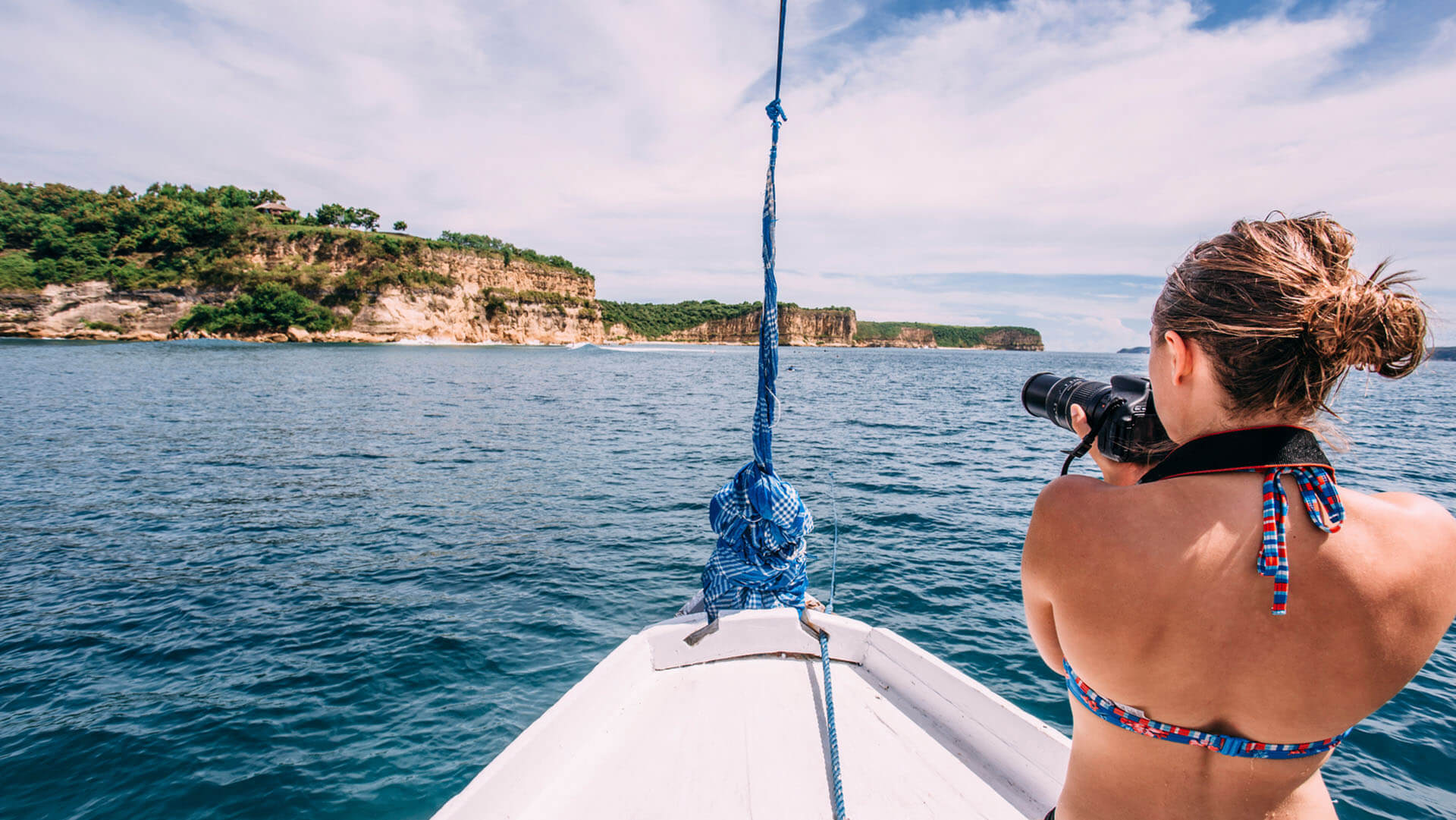 Blond girl in bikini on boat at sea taking photograph of ocean landscape
