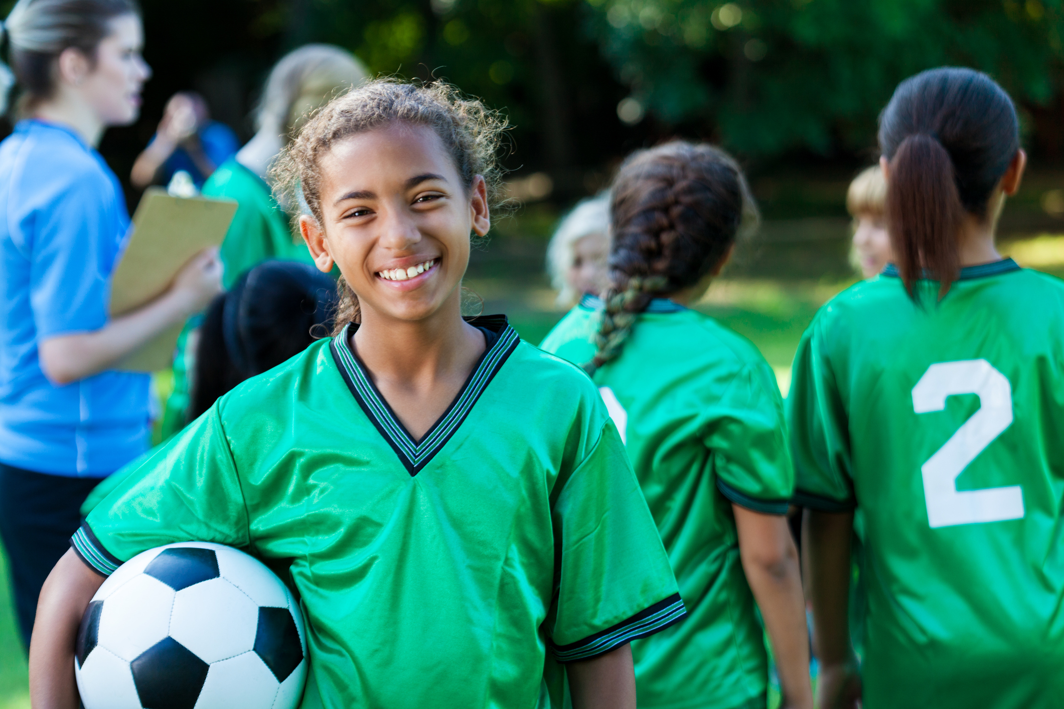 Pretty preteen soccer player smiles at the camera while holding a soccer ball. She is wearing a green jersey. Her team and coach are in the background.