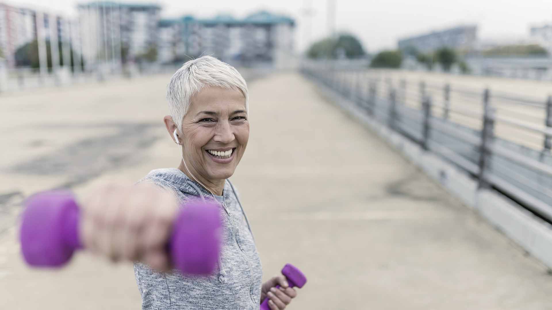 873945600 Portrait of cheerful aged woman Fighting breast cancer in fitness wear exercising with pink dumbbells in the city during cloudy day. Front view of Happy Gray hair female doing dumbbell curls in the fresh air. Smiling, Joyful senior, short hair, lady exercising with weights outdoors. Health and sport.