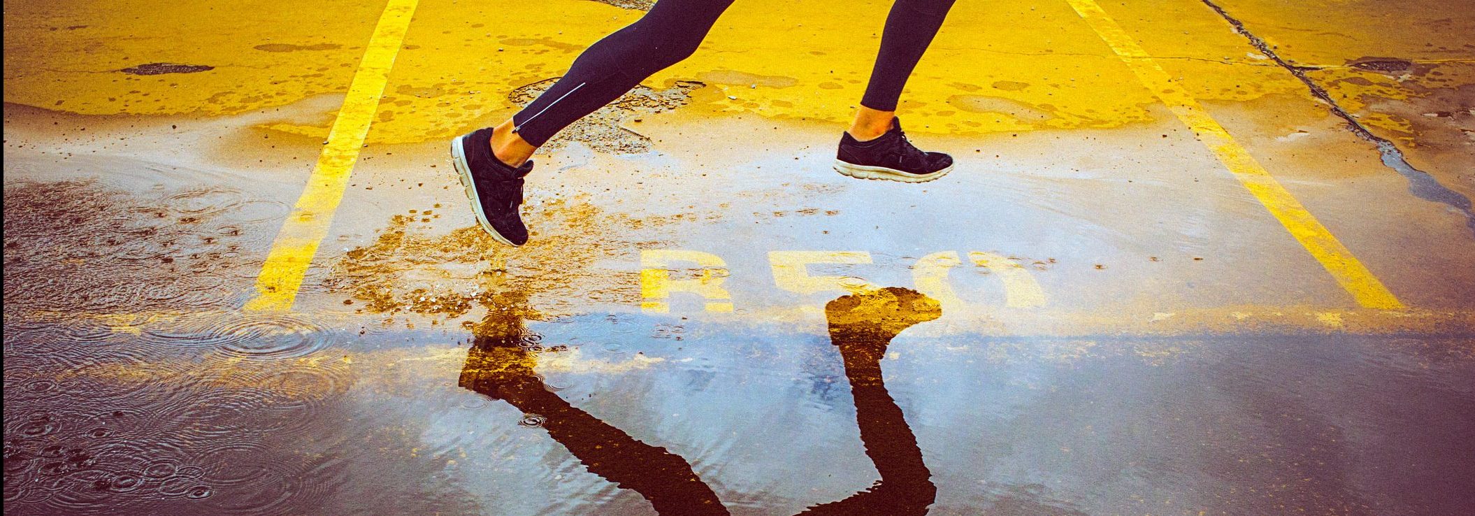 Young person running over the yellow parking lot. Black sport clothing - sport shoes, running tights, and a jacket. High angle view of a runner's legs and its reflection in the water.