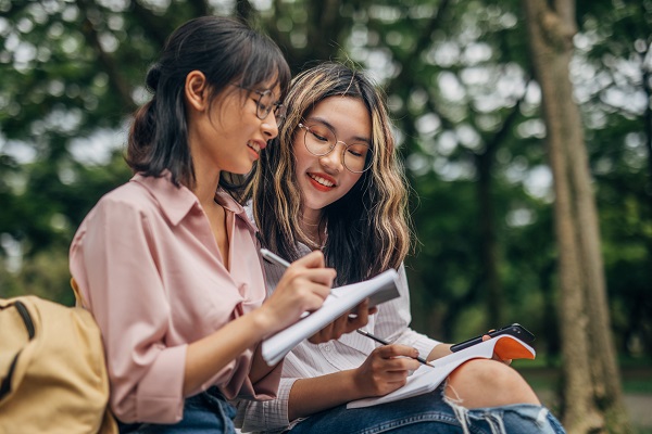 Two Taiwanese female students with backpacks sitting outdoors and writing homework.