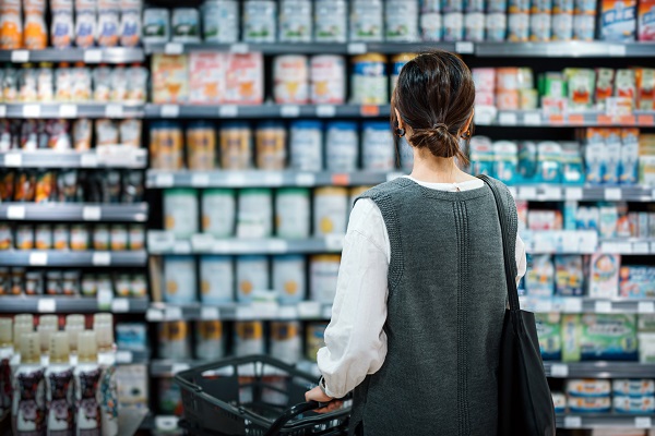 Rear view of young Asian mother with a shopping cart grocery shopping for baby products in a supermarket. She is standing in front of the baby product aisle and have no idea which product to choose from