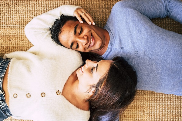 Young pretty interracial couple lay down on the carpet and smile having care and love of each other - happy and cheerful above portrait of boy and girl enjoying home together - concept of life together