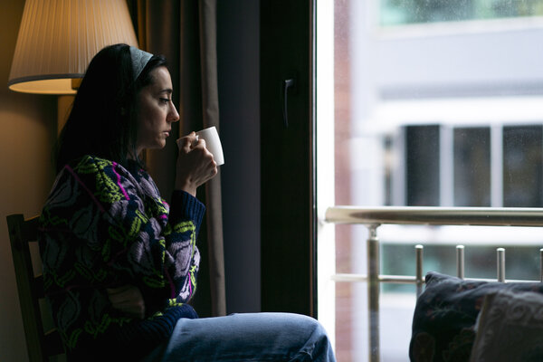 Sad woman sitting on a sofa, drinking a cup of coffee and looking trough the window at home.