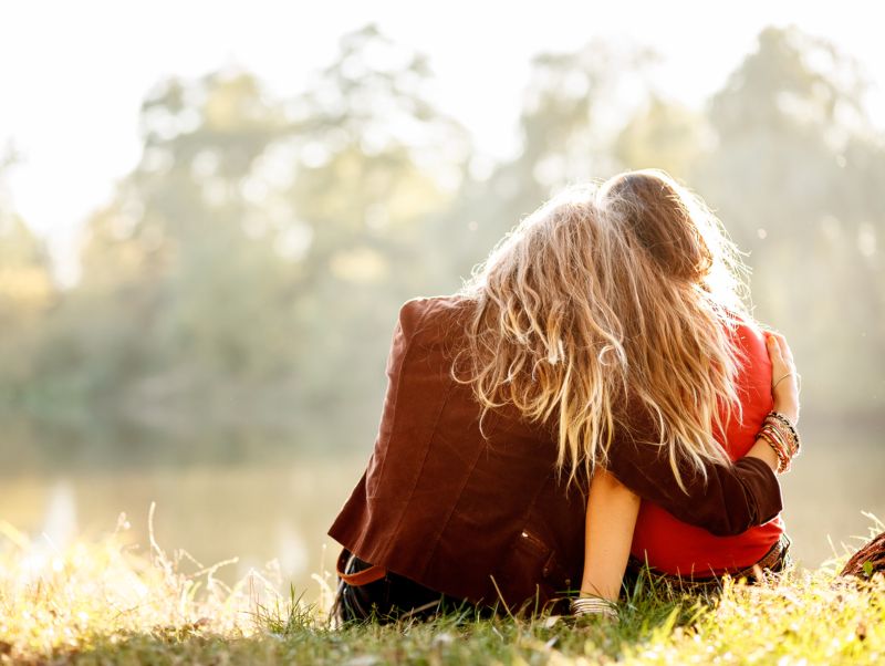 two young women sitting on grass hugging rear view