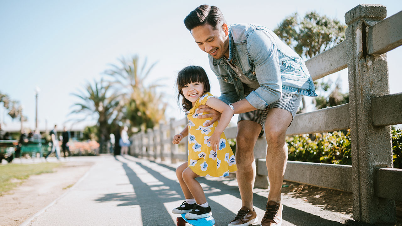father helping young daughter to skateboard on a footpath